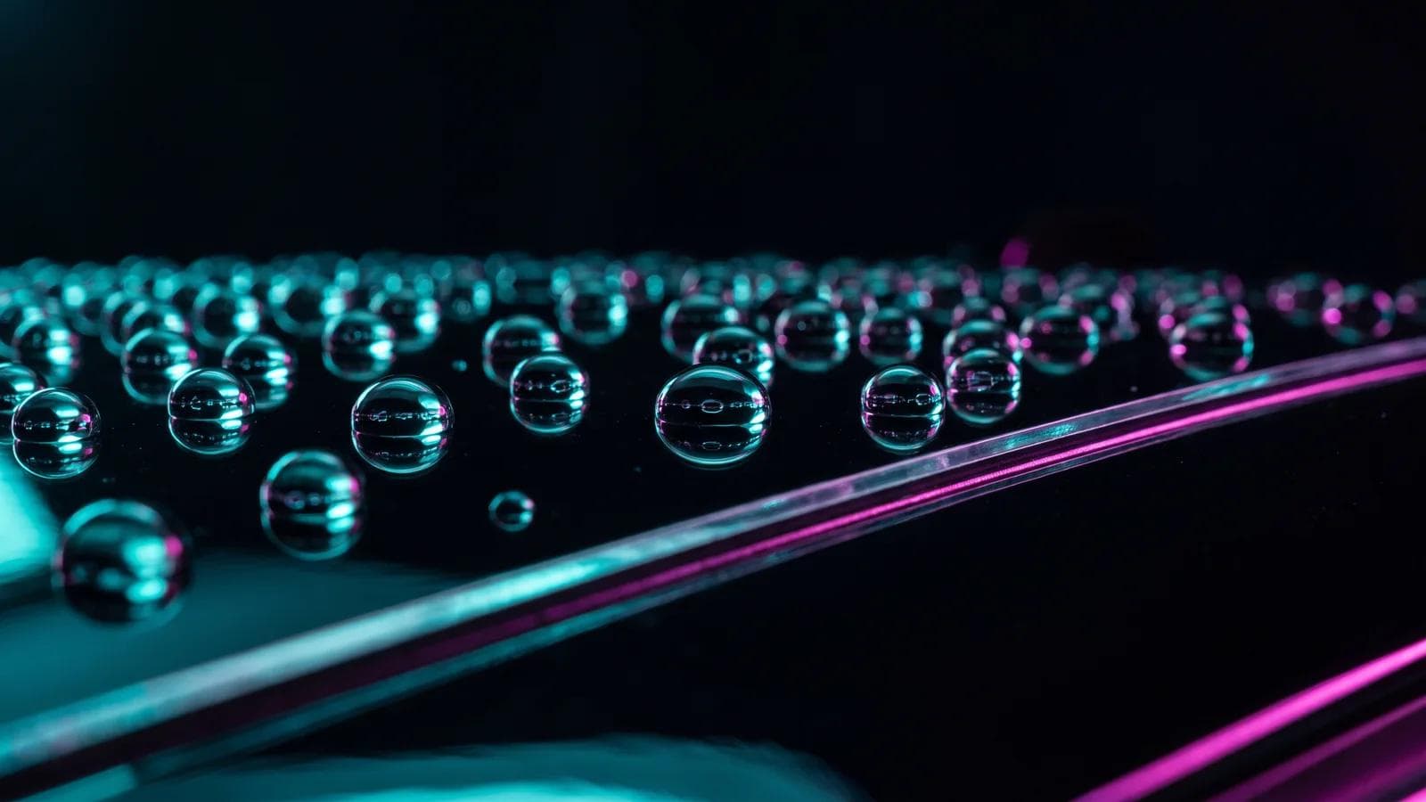 Perlende Wassertropfen auf tiefschwarzem, keramikversiegeltem Autolack neben einer Folienkante, mit türkis-magentafarbenen Neonreflexen.